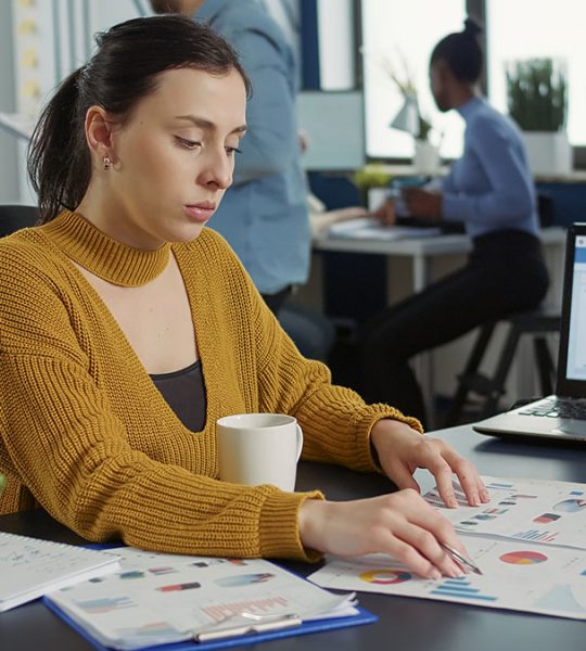 Portrait of startup employee comparing two sheets of paper with charts analyzing financial data while drinking morning coffee. Woman working in busy marketing department sitting at desk with laptop.